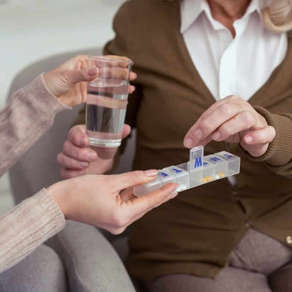 Home Care Services - Medication Reminders. Close up of attractive female hand holding container for medication while giving glass with water to elder woman