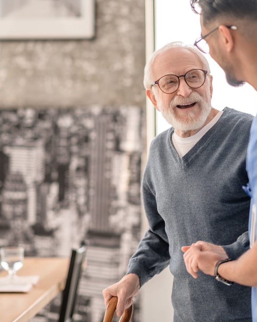 About Ally In-Care. Old man standing by the table supported by a male nurse