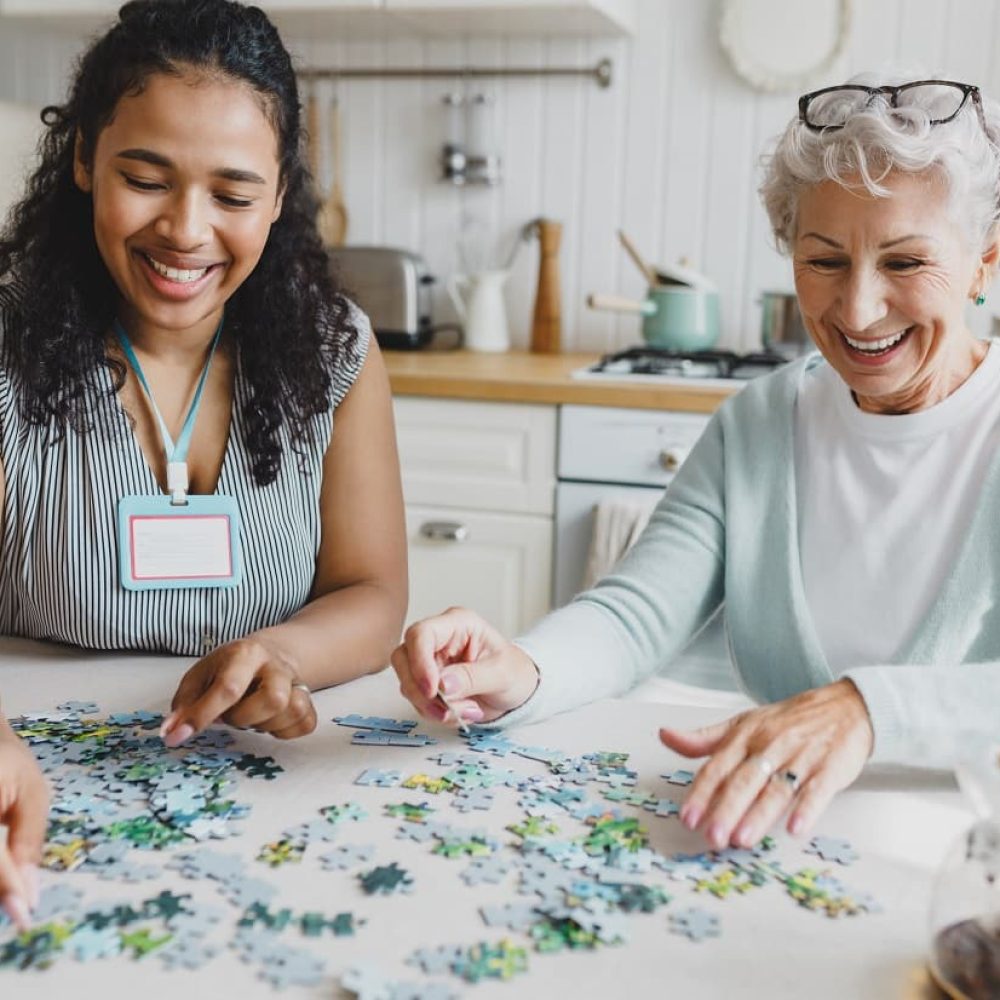 home care services - Companionship. Portrait of african american female social worker volunteer playing puzzle game with cheerful senior lady, having fun, laughing searching for right piece sitting together at kitchen table